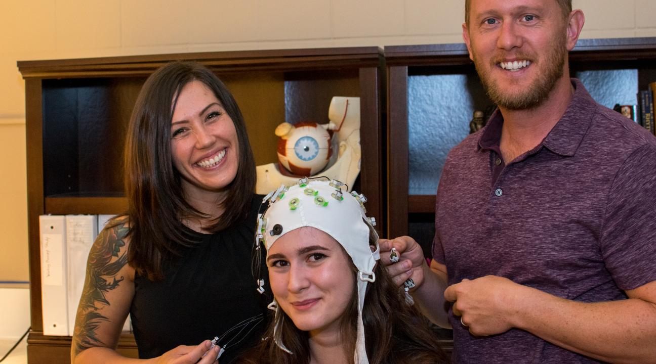 Student Olivia Krieger, left, and Professor Jesse Bengson demonstrate a cap used to record brainwaves with student Alexandra Theodoroub