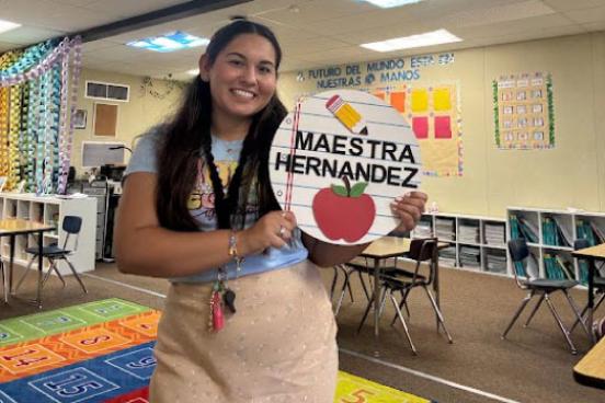 Nataly Hernandez, an SSU alumna, is now a 2nd grade teacher in Winters, California. In this image she is standing in her classroom holding a sign that says "Maestra Hernandez."