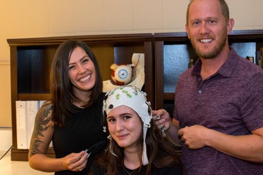 Student Olivia Krieger, left, and Professor Jesse Bengson demonstrate a cap used to record brainwaves with student Alexandra Theodoroub