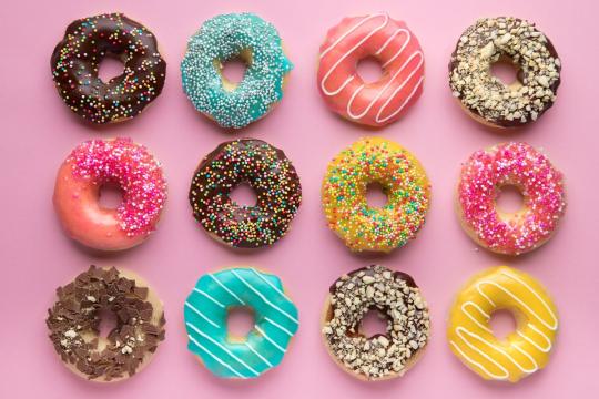 A variety of twelve different colored/flavored donuts in front of a pink background