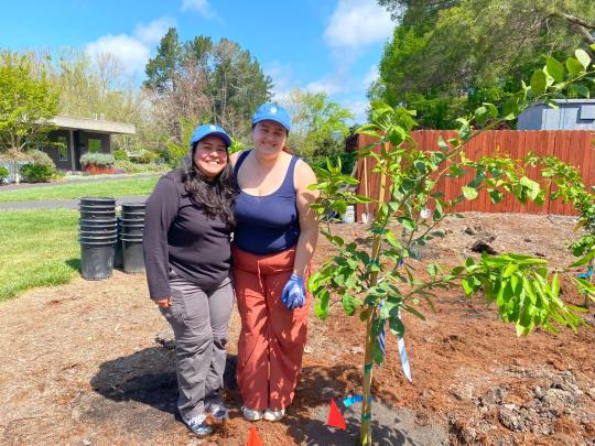 Two people smiling and posing next to a freshly planted tree